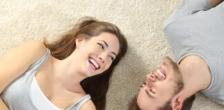 Couple laying on their back with heads turned towards each other and smiling widely on a beige carpet for relationship green flags