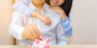 Smile couple putting a coin into a pink piggy bank on wooden desk - save money for the future