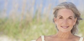 An attractive elegant senior woman sitting on a white sand beach with grass and a blue sky behind her.