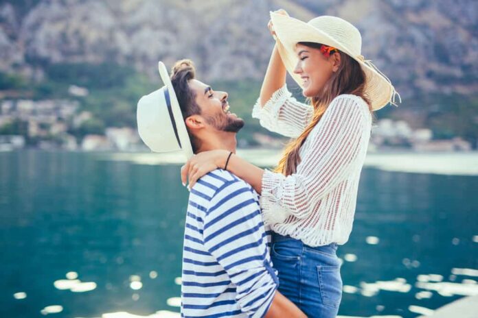 long distance date ideas header - couple in front of mediterranean water smiling and laughing. man is lifting up girl, both are wearing white hats