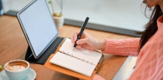 woman in pink sweater writing in a notebook in front of a computer 