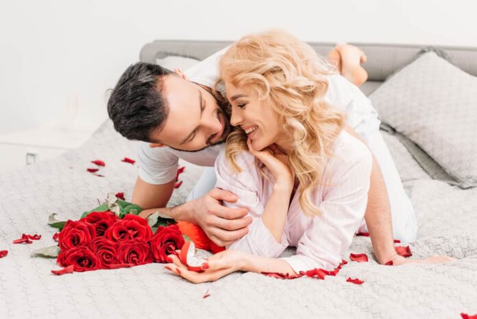 valentine's day date ideas - couple cuddling on bed next to bright red bouquet of roses