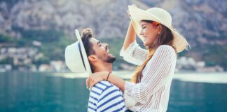 honeymoon in europe - couple laughing next to a mediterranean coast. both are in white and blue wearing summer hats