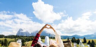 A couple lays on their backs in a field while making a heart with their hands, planning a romantic getaway together