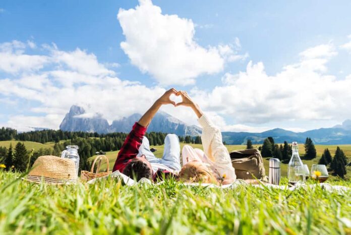 A couple lays on their backs in a field while making a heart with their hands, planning a romantic getaway together