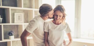 A man kisses his spouse on the cheek as she chops vegetables in a sunny kitchen while discussing how to manage expectations in marriage