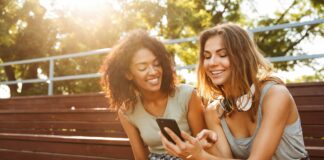 how to manifest a text message - two women sit outside at a bench looking at a phone together and smiling